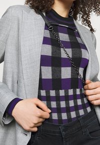 Veste grise portée sur un pull à carreaux noir et violet avec col côtelé noir. Le mannequin tient une bandoulière en chaîne, mettant en valeur un jean noir ajusté.