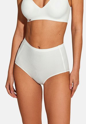 Femme portant des sous-vêtements côtelés blancs taille haute et un bralette assorti, debout les bras détendus le long du corps sur un fond uni.