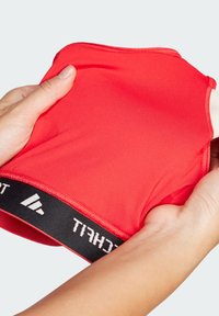 Top deportivo atlético rojo sostenido con las manos, presenta una textura suave, escote redondeado y una banda elástica negra con marca y letra blanca.