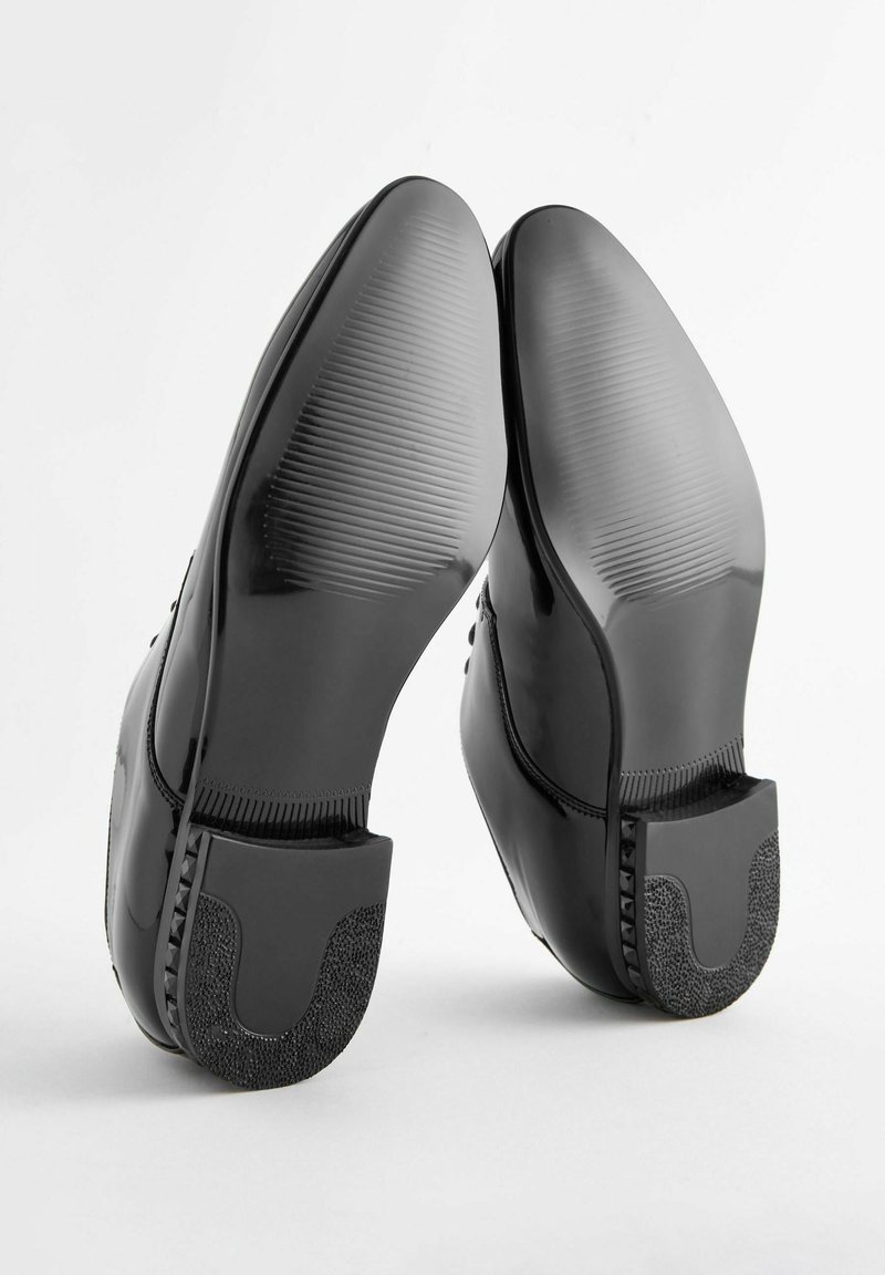 Schwarze, glänzende Schuhe mit abgerundeten Zehen, strukturierten Gummisohlen und einem gemusterten Absatzakzent. Glatte Oberfläche mit klaren Linien und minimalen Details.