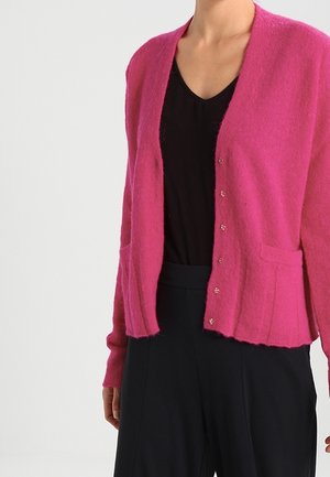 Donna che indossa un cardigan rosa acceso con bottoni sopra una maglia nera a scollo a V e pantaloni neri, su uno sfondo chiaro e uniforme.