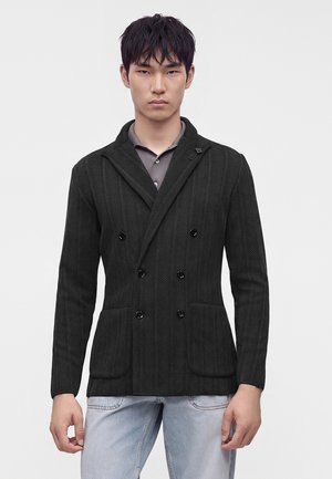 Junger Mann trägt einen dunklen, doppelt geknöpften Blazer mit Fischgrätmuster über einem grauen Hemd und hellblauen Jeans vor einem schlichten weißen Hintergrund.