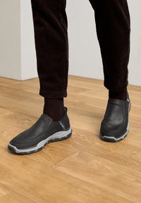Black slip-on shoes with textured leather uppers and a gray rubber outsole, set against a wooden floor and corduroy pants.