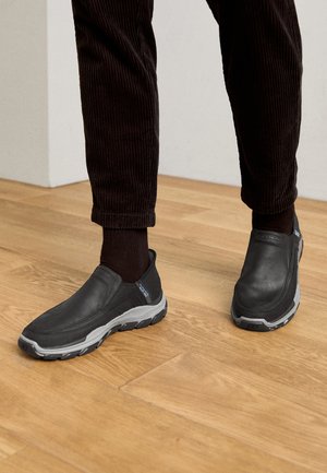 Sorte slip-on sko med strukturerede læderoverdele og en grå gummi sål, placeret på et trægulv og med fløjlsshorts.