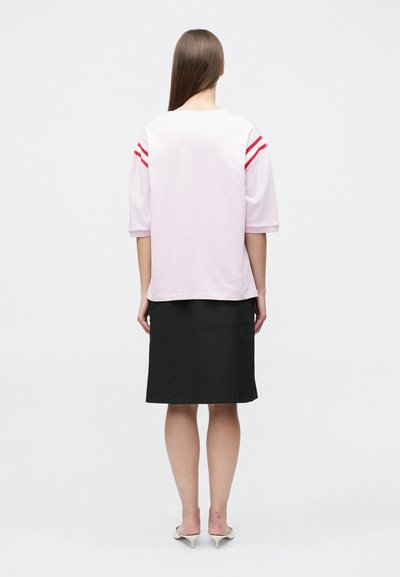 Γυναίκα με μακριά ίσια μαλλιά που φοράει ροζ ανοιχτό πουκάμισο με κόκκινες ρίγες στα μανίκια, μαύρη φούστα μέχρι τα γόνατα και λευκά παπούτσια με τακούνι, γυρισμένη πλάτη.