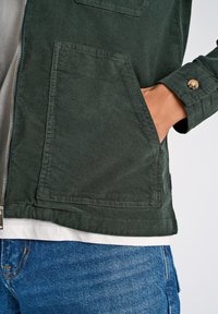 Groene corduroy jas met rits, twee voorzakken en een knoopmanchet. Gdragen over een lichtgrijze shirt en blauwe spijkerbroek.