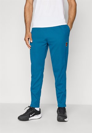 Mand iført teal atletisk bukser med et patch på venstre lår, sort og hvid Nike sneakers og en hvid T-shirt med korte ærmer.