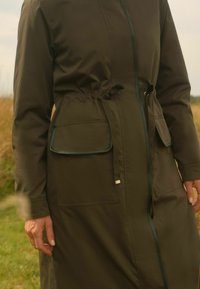 Olivově zelený dlouhý kabát s šňůrkou v pase, dvěma předními kapsami a kontrastním lemem. Hladká textura, funkční design vhodný pro nošení venku.