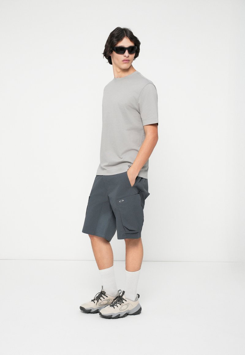 Graues T-Shirt mit kurzen Ärmeln, dunkelgraue Cargo-Shorts mit Seitentaschen, kombiniert mit beigen Schuhen und weißen Kniestrümpfen sowie schwarzen Sonnenbrillen.