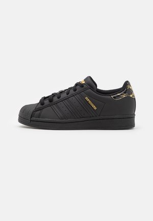 Zapatilla negra con parte superior de cuero, suela de goma, puntera redondeada, tres rayas laterales, detalle dorado y tacón decorativo en negro y dorado.