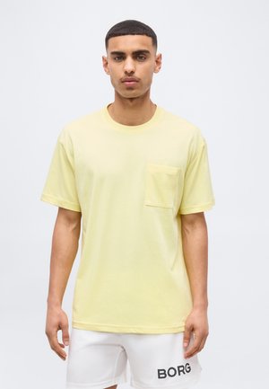Άνδρας με κοντά μαλλιά που φοράει ανοιχτό κίτρινο μπλουζάκι με τσέπη στο στήθος και λευκό σορτς με το λογότυπο «BORG», στέκεται μπροστά σε απλό φόντο.