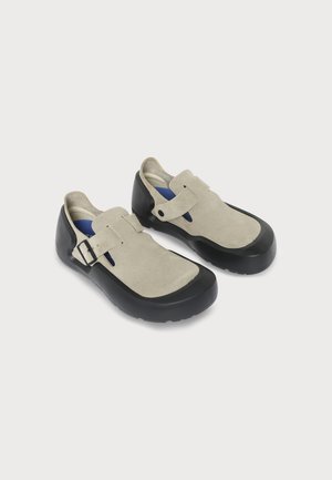 Paire de chaussures en daim beige avec semelles en caoutchouc noir et brides latérales réglables sur un fond clair uni.