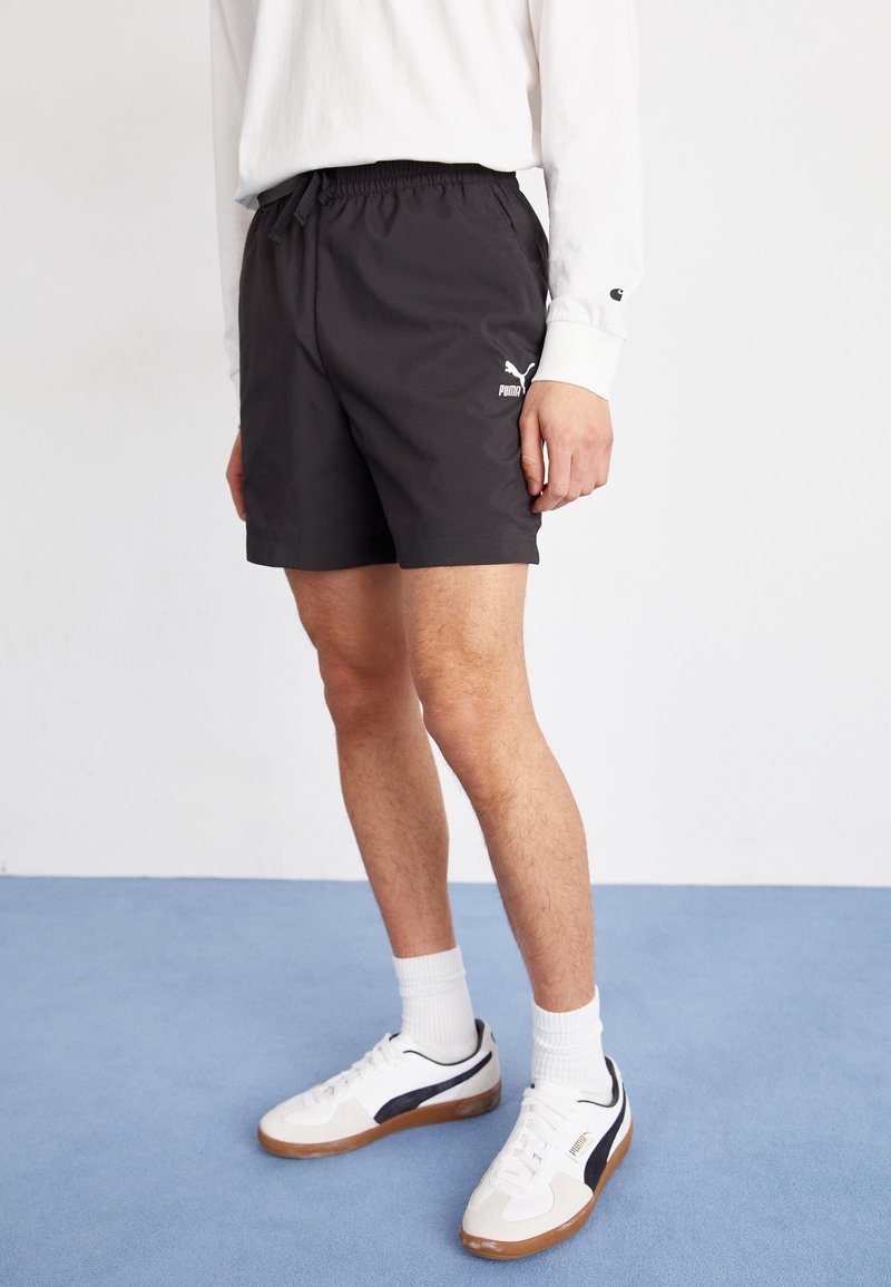 Homme portant un short noir Puma, une chemise à manches longues blanche, des baskets blanches Puma et des chaussettes blanches, debout sur un sol bleu.