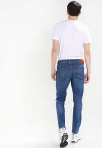 Smal blå jeans med en slät textur, femficksdesign och en tan läderlapp på baksidan av midjan. Bärs med en vit t-shirt och sneakers.