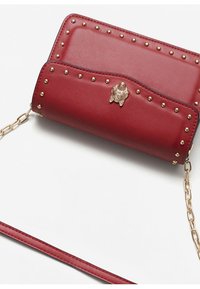 Červená kožená crossbody taška se zlatými hroty, texturovaným klopou a dekorativním zlatým emblémem. Obsahuje řetízkový popruh.