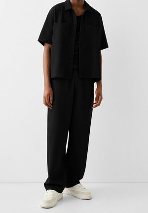 Personne portant une chemise noire ample à manches courtes boutonnée et un pantalon large assorti avec des baskets blanches, debout devant un fond uni.