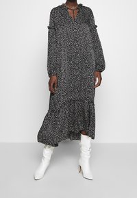 Robe noire à manches longues avec des pois blancs, ourlet à volants et décolleté en V avec un détail à nouer, portée avec des bottes blanches jusqu'aux genoux.
