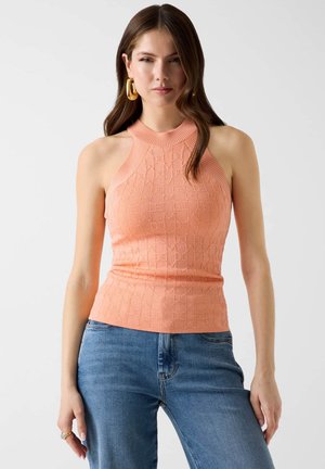 Femme portant un haut sans manches à motifs couleur pêche et un jean bleu, avec de longs cheveux bruns et de grandes boucles d'oreilles en or.