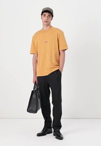 Gul bomulds T-shirt med rund hals, sorte bukser, sorte sko og en mønstret kasket. Holder en sort lædertaske.
