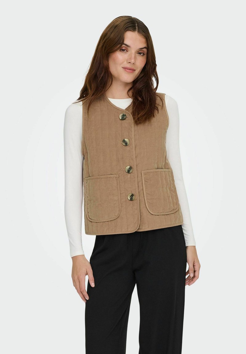 Donna che indossa un gilet trapuntato beige con bottoni sopra una maglia bianca a maniche lunghe e pantaloni neri, in piedi davanti a uno sfondo chiaro e neutro.