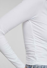 Weißes Langarmshirt aus dehnbarem Baumwollstoff, mit Seitennähten und taillierter Silhouette. Glatte Textur mit leichtem Glanz.