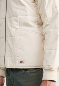 Beige pufferjakke med quiltet design, der har lynlåslukning foran og en sidelomme. Ribstrikkede manchetter og et lille logo-patch.