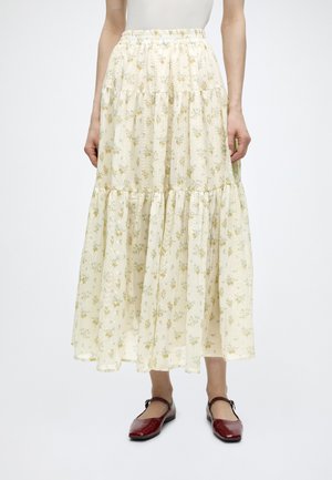 Kobieta ubrana w kremową, warstwową kwiatową spódnicę z elastycznym pasem i czerwone płaskie buty typu Mary Jane.