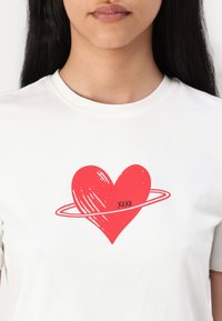 Balta crew-neck marškinėliai su raudonu širdies grafiku, apsuptu plonos raudonos linijos, viduje širdies yra užrašas "XOXO".
