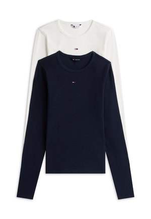 Junge Frau trägt ein weißes Langarmshirt, blaue weit geschnittene Jeans, Sneakers und einen lila-weißen Schal über den Schultern.