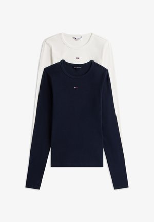 Młoda kobieta ubrana w biały top z długim rękawem, niebieskie spodnie typu wide leg, sneakersy i fioletowo-biały szalik przewieszony przez ramiona.