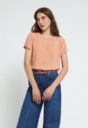Femme aux longs cheveux bruns portant un chemisier pêche à manches courtes, un jean bleu taille haute à jambes larges, et une ceinture marron avec une boucle dorée.