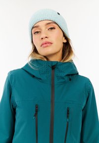 Türkise wasserdichte Jacke mit einer enganliegenden Kapuze, schwarzen Reißverschlüssen und perforierten Belüftungsdetails. Kombiniert mit einer hellblauen gerippten Mütze.