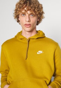 Hoodie amarelo de algodão com capuz ajustável, bolso canguru e logótipo da Nike em branco no peito. Tecido texturizado com um acabamento suave.