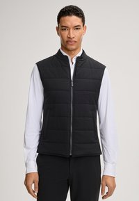 Schwarze gesteppte Weste mit Reißverschluss vorne, zwei Seitentaschen und hohem Kragen; getragen über einem weißen Langarmshirt. Glatte Stoffoberfläche.