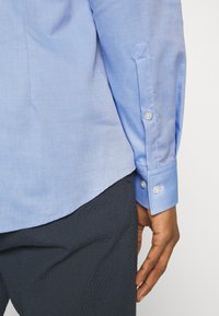 Světle modrá košile s texturovanou látkou, zdobená manžetou na knoflíky a detailem postranního švu, doplněná tmavými kalhotami s texturou.