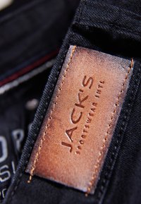 Tissu en denim noir avec une étiquette en cuir marron embossée avec "JACK'S" et des détails de couture. Gros plan sur l'étiquette de vêtement.