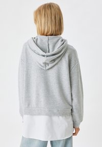 Sudadera con capucha gris de textura suave, diseño corto y corte holgado. Presenta un bolsillo tipo canguro y puños de canalé, llevada sobre una camiseta blanca.