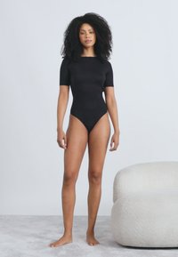 Μαύρο κορμάκι με κοντά μανίκια και ομαλή υφή, με εφαρμοστή σχεδίαση και ψηλό λαιμό, συνδυασμένο με ένα ανοιχτόχρωμο, στρογγυλεμένο καναπέ.