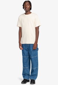 T-shirt di cotone color crema con maniche corte, abbinato a pantaloni in denim blu a gamba larga con cuciture in contrasto e tasche. Scarpe nere.