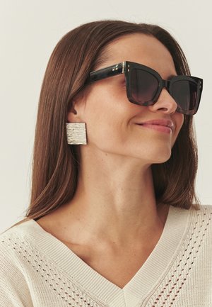 Egyenes barna hajú nő, nagy, négyzet alakú fekete napszemüveget, texturált, téglalap alakú ezüst fülbevalókat visel, valamint krémszínű kötött V-nyakú felsőt.
