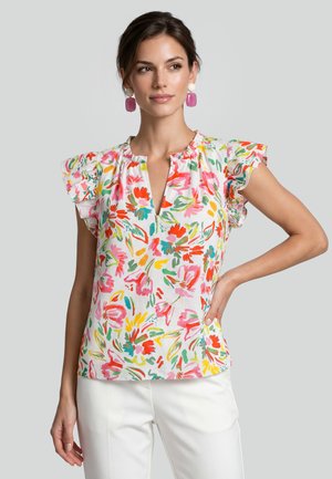 Kvinde iført en farverig blomsterbluse med flæseærmer og hvide bukser, poserende med den ene hånd på hoften mod en ensfarvet baggrund.