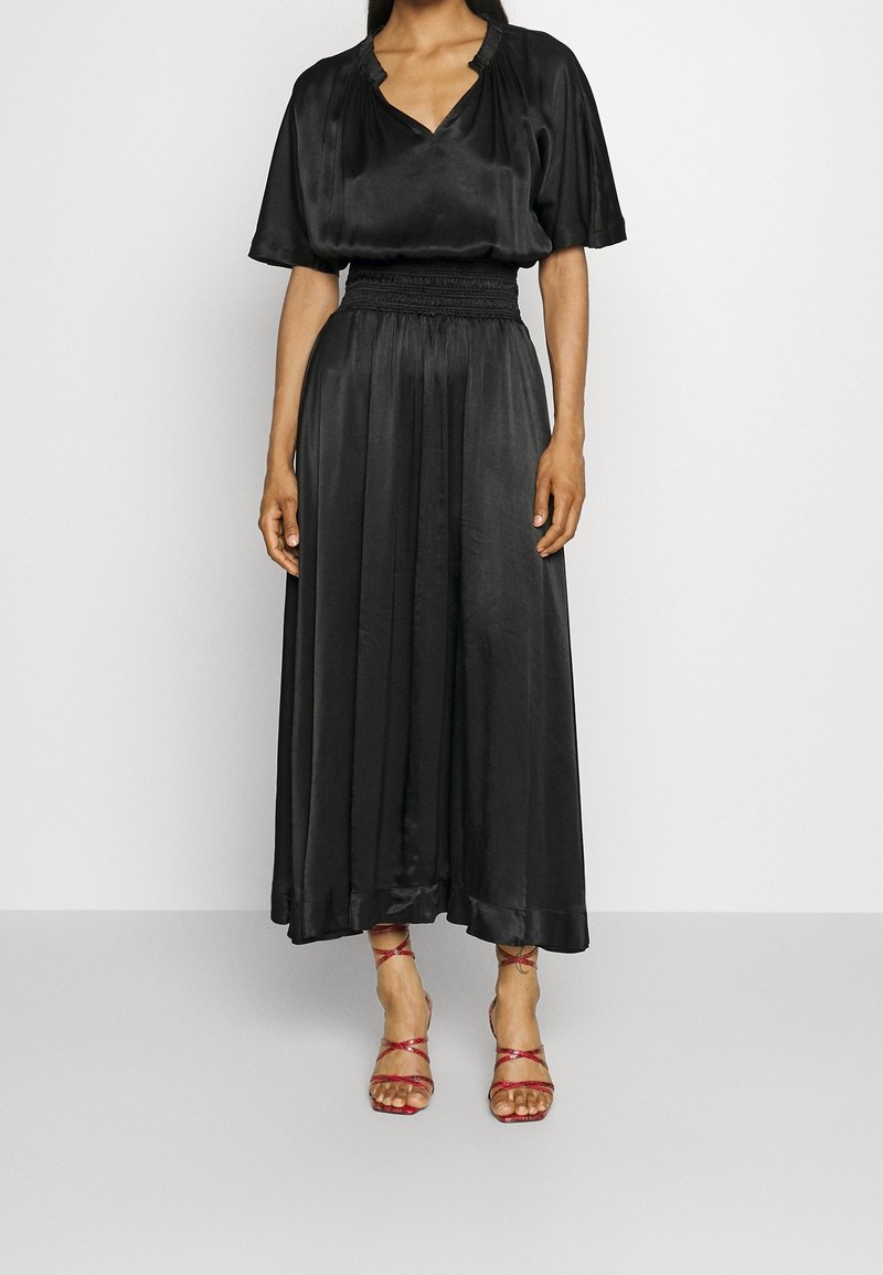 Femme portant une robe midi noire à manches courtes et col en V, avec une taille froncée et des sandales rouges à talons hauts et fines brides, debout contre un mur blanc.