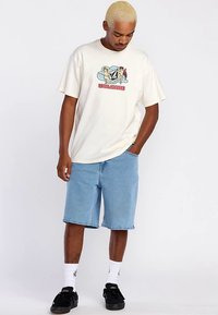 T-shirt crème à manches courtes avec un imprimé graphique, associé à un short en denim bleu clair et des baskets noires, avec des chaussettes blanches chevilles.