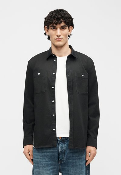 Jeune homme aux cheveux bouclés portant une chemise noire boutonnée sur un t-shirt blanc et un jean bleu, debout devant un fond clair uni.