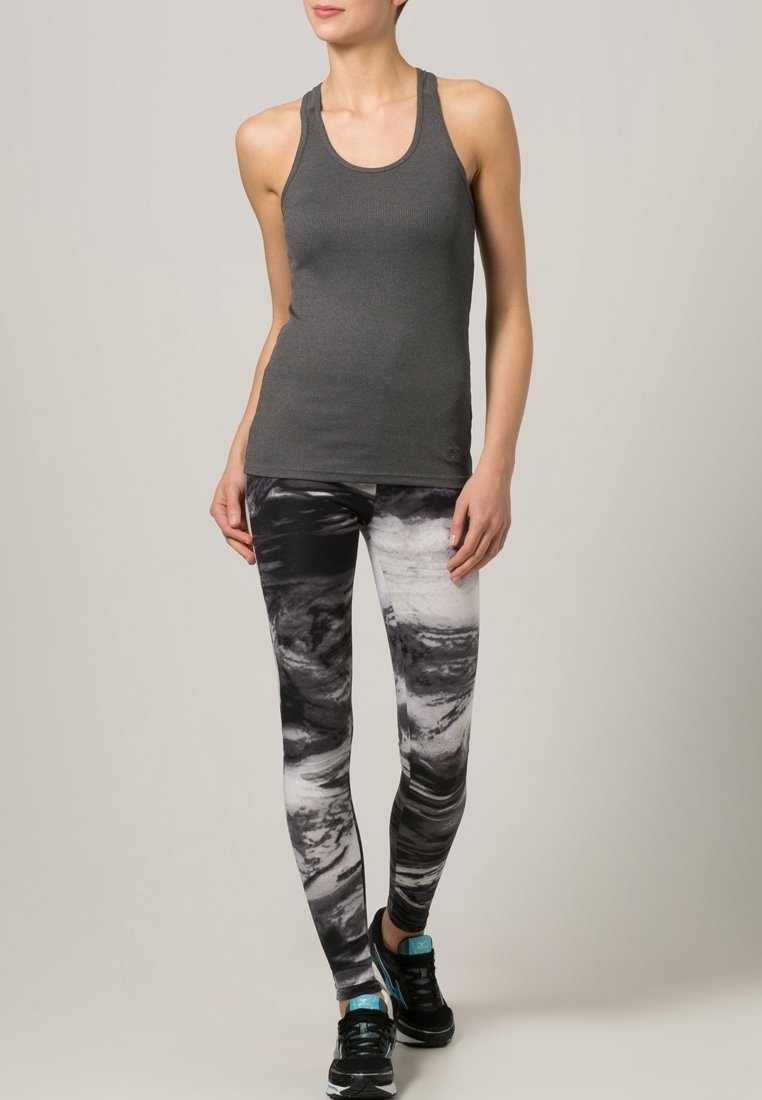 Femme debout portant un débardeur gris foncé, des leggings à motifs noirs et blancs, et des baskets noires avec des touches bleues sur un fond uni.
