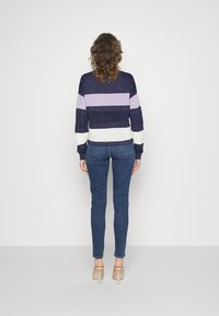 Persona con capelli ricci che indossa un maglione a righe blu navy, lavanda e bianco, jeans skinny blu e sandali con tacco trasparente, di spalle.
