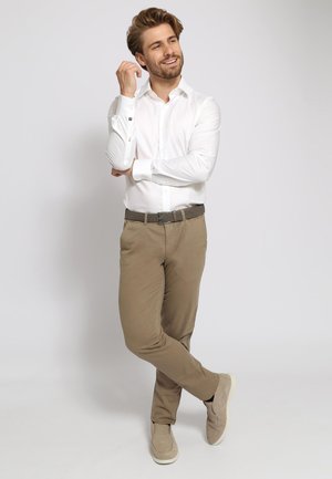Witte lange mouw shirt, kaki broek en lichtgekleurde instap schoenen. Heeft een geweven bruine riem en een gladde stofstructuur.