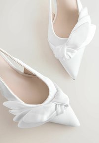 Chaussures slingback blanches à bout pointu avec des détails en tissu noués semblables à des ailes à l'avant, sur un fond clair.