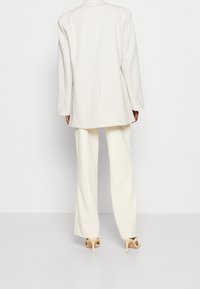 Bílý oversize blejzr s strukturovaným designem, s jedním rozparkem a dlouhými rukávy, doplněný širokými bílými kalhotami a tělovými lodičkami.