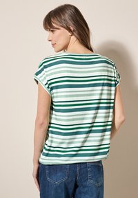 Top de manga corta a rayas verdes y blancas, hecho de un tejido suave, que presenta un corte relajado y un cuello redondeado. Combinado con jeans de mezclilla azules.